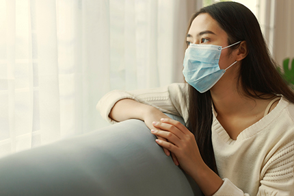 Young Asian woman wearing protective face mask stay quarantine at her apartment. Lonely women sitting on a couch looking out of the window.